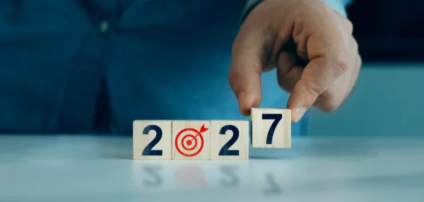 A person’s hand changes a wooden block with the number 6 to 7, forming “2027.” The middle block shows a target with an arrow. The background is blurred, focusing on the blocks and hand.