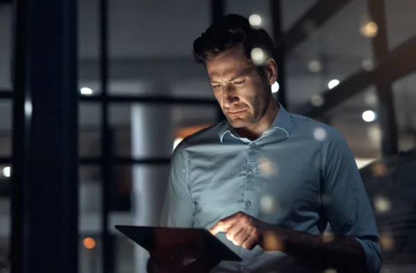 A man in a light blue shirt stands in a dimly lit office at night, looking down and using a tablet, with reflections of lights on the glass—perhaps reviewing Ontario Compensation Benchmark data to ensure pay transparency.