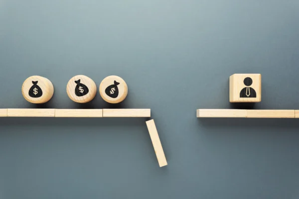 Wooden blocks with money bag symbols on one side of a gap, a block falls between, and a block with a person icon sits on the other side, symbolizing a compensation problem or barrier between employee pay and the individual.