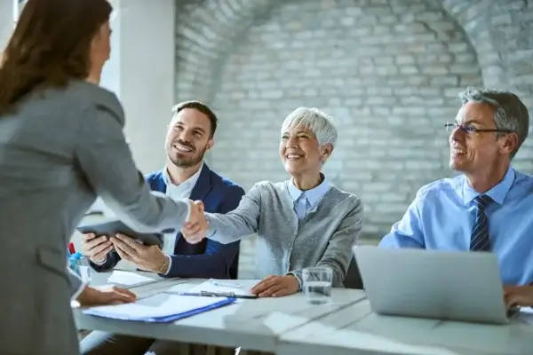 Four professionals in business attire sit at a table during a meeting; one woman stands and shakes hands with a smiling woman across the table, while two men watch. Ontario employers discuss competitive pay, with a laptop and documents on the table.