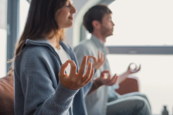 Two people sit indoors in a meditative pose with eyes closed and hands resting on their knees, making a mudra gesture. The focus is on their hands, creating a calm and peaceful atmosphere that echoes the growing emphasis on workplace wellness initiatives in Ontario.