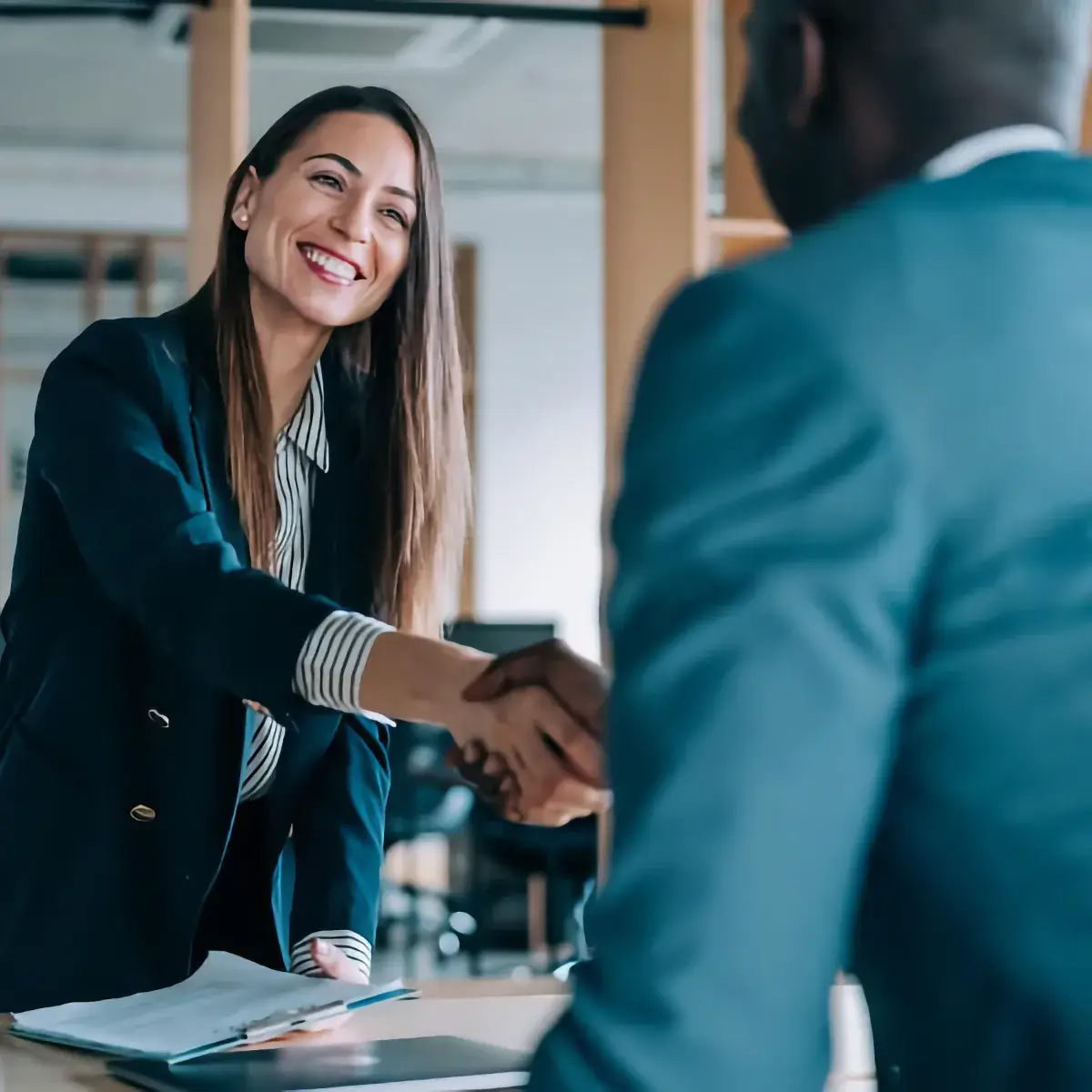 A woman in business attire smiles and shakes hands with a man across a desk in an office setting, suggesting a successful meeting discussing compensation management. A clipboard and papers are visible on the desk.