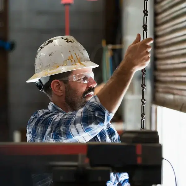 A man wearing a white hard hat, safety glasses, and a plaid shirt pulls on a chain to open or close a metal industrial garage door inside a workshop where employee compensation is carefully considered.