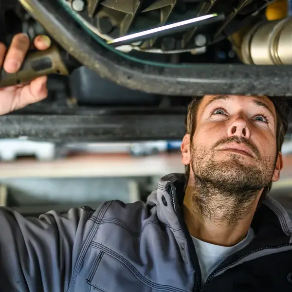 A man wearing a gray jacket inspects the underside of a vehicle, looking upward while holding a tool in his hand—illustrating the hands-on side of salary benchmarking in technical roles.