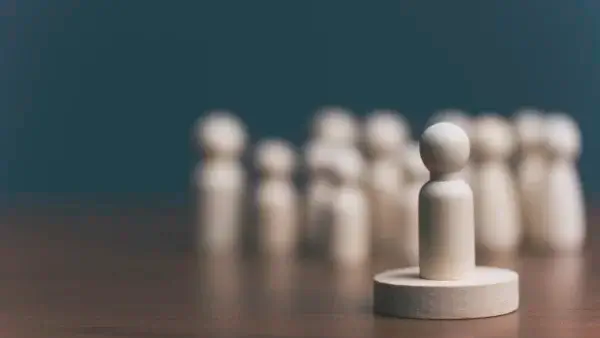 A wooden figure stands on a round platform in the foreground, with several similar figures blurred in the background, symbolizing leadership or individuality.