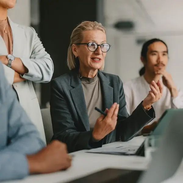 A woman with glasses discusses compensation management during a meeting, gesturing as two colleagues listen attentively beside her in a modern office setting.