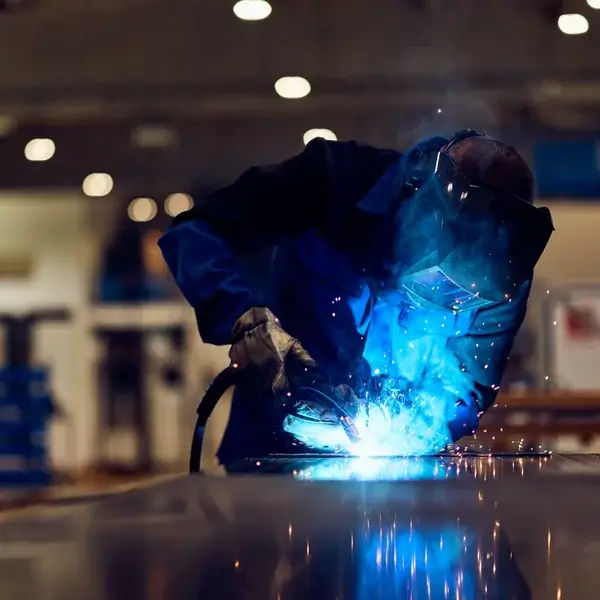 A person wearing protective gear and a welding helmet uses a welding torch, producing bright blue sparks in an industrial workshop where employee compensation and safety are top priorities.